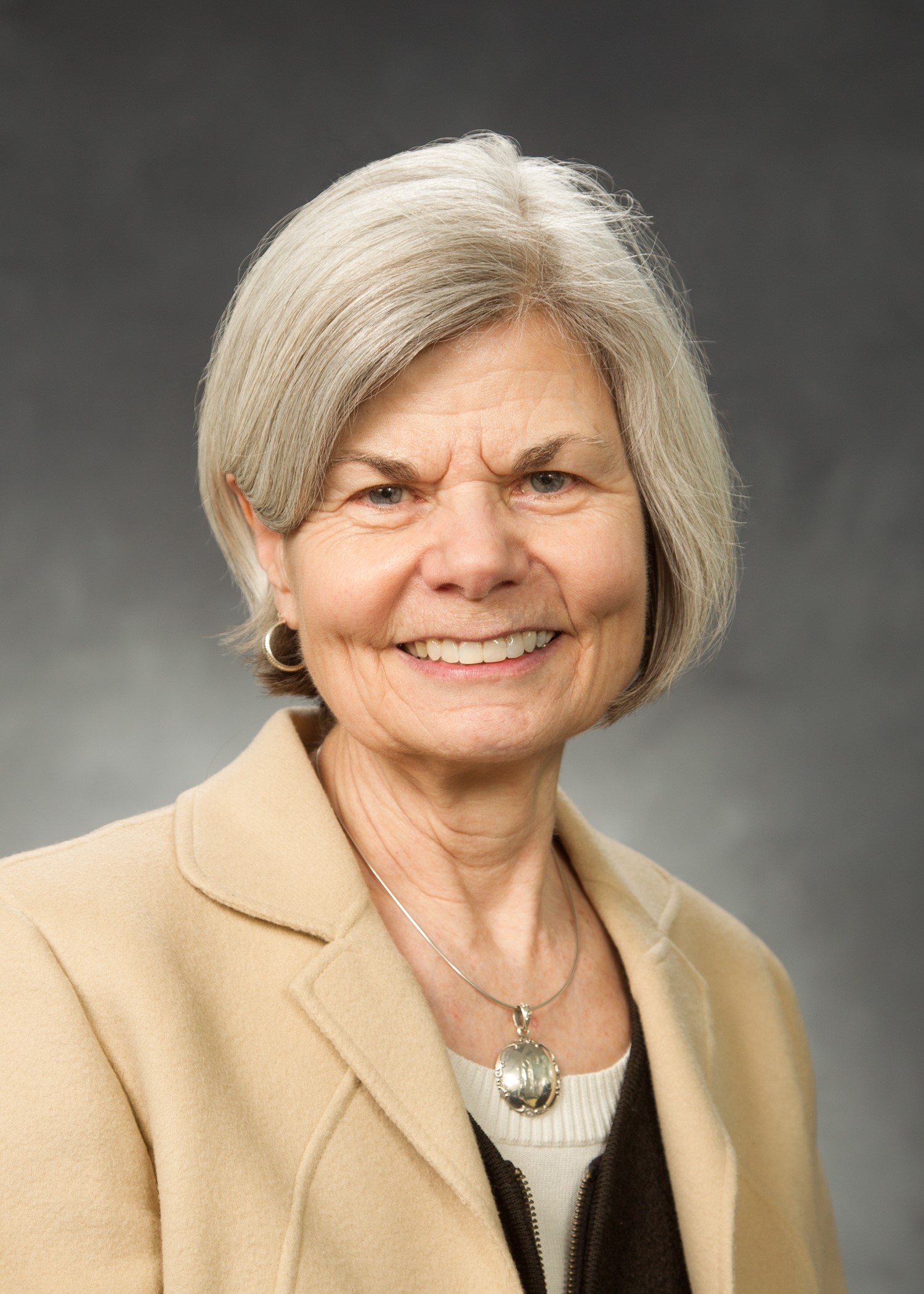 An older adult with short gray hair smiles in a beige blazer and necklace against a gray background.