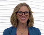 A person with shoulder-length hair and glasses smiles in a blue jacket against a textured white backdrop for medbridge.