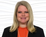 A person with long blonde hair in an orange top, black blazer, and pearl necklace stands against a white background, representing medbridge's role in digital healthcare integration.