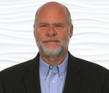 A bearded man in a blue shirt and dark blazer stands against a wavy white background, representing digital healthcare innovation at medbridge.