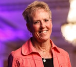 Smiling woman with short gray hair in a coral jacket, in front of a purple-lit modern medbridge digital healthcare background.
