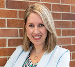A woman with blonde hair wearing a blazer stands smiling against a brick wall.