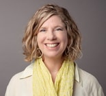 Smiling woman with wavy blonde hair, pale shirt, and yellow scarf poses in front of a gray background.