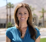 Smiling woman with long brown hair and blue blouse stands outside with mountains and palm trees, representing medbridge healthcare.