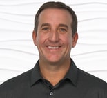 A person in a dark polo shirt smiles against a wavy white background.