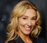 Smiling blonde woman in a dark blazer on a dark background.