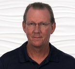 A middle-aged person with glasses and tied-back hair in a dark shirt stands against a white background, reflecting digital healthcare themes relevant to medbridge.