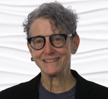 A smiling person with gray hair and glasses wears a black jacket and blue shirt against a white textured background.