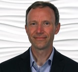 A man in a suit jacket and collared shirt smiles before a wavy white background for medbridge.