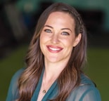 A smiling woman with a braid, in a teal top and gold necklace, stands before a blurred green background for medbridge.