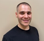 Smiling person with short hair and trimmed facial hair in a black t-shirt stands in front of a beige wall, suitable for medbridge digital healthcare.