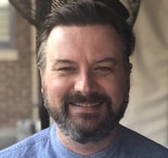 Man in a light blue shirt smiles against a blurred outdoor background, suggesting his role in healthcare education or medbridge software.
