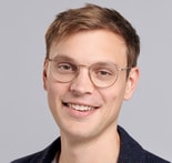 A young man with short light brown hair, round glasses, and a dark jacket smiles at the camera against a light gray background, representing medbridge.