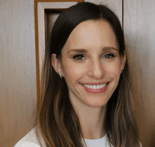 A woman with long brown hair in a white top smiles at the camera in front of a wooden wall, representing medbridge warmth.