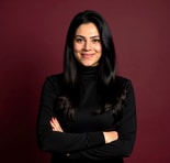 Woman with long dark hair in a black turtleneck smiles confidently with arms crossed before a maroon background, for medbridge.