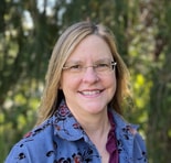 A woman with long blonde hair and glasses smiles outdoors, wearing a blue floral shirt and maroon top, with greenery in the background.