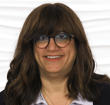 A smiling person with long brown hair and glasses, wearing a blazer and collared shirt, stands in front of a white textured background.