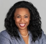 Woman with long curly hair, grey blazer, and pearl necklace smiles at camera.
