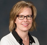 A smiling person with short blonde hair and glasses, wearing a white sweater over a black floral-patterned shirt, embodies their passion for digital healthcare innovations at medbridge.