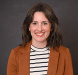 Smiling woman with shoulder-length brown hair in a brown blazer and striped shirt, in front of a dark background.