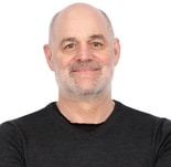 A middle-aged, bald man with a gray beard and dark sweater smiles against a white background, representing medbridge.