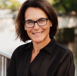 A woman with short brown hair and glasses smiles outdoors by water, representing positive medbridge healthcare training.