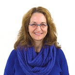 A woman with long, wavy hair and glasses smiles warmly in a blue knitted sweater and scarf, embodying medbridge's digital healthcare innovation.