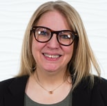 A smiling person with long blonde hair, glasses, and a black blazer stands against a white wavy background.