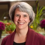 A smiling woman with short gray hair in a burgundy blazer stands before blurred green plants and purple flowers at medbridge.