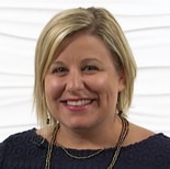 A smiling person with short blonde hair, in a navy blue top and beaded necklace, embodies professionalism in Medbridge healthcare training.