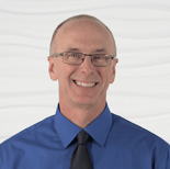 A person wearing glasses and a blue shirt smiles against a wavy white backdrop.