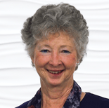 Smiling elderly woman with gray hair, in a dark top and floral scarf, against a textured white background.