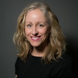 Smiling blonde woman in a black top and hoop earrings against a dark gray background.