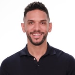 A smiling bearded person in a dark shirt stands against a plain white background.