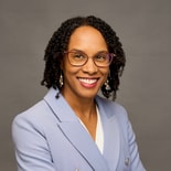 A person with curly hair, glasses, and a blue blazer smiles against a gray background, representing Medbridge in digital healthcare.