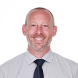 A smiling bearded man in a light patterned shirt and dark tie stands against a white background, symbolizing medbridge's integration into patient care.
