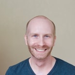 A bald man with a beard smiles in a dark V-neck shirt against a plain background.