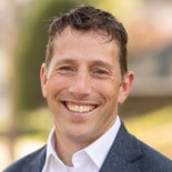 A man in a dark blazer smiles outdoors with greenery, representing medbridge healthcare education.