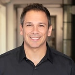 A man with short dark hair in a black collared shirt smiles indoors, representing medbridge healthcare education.