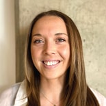 A woman with straight brown hair smiles at the camera in front of a concrete wall, representing medbridge healthcare training software.