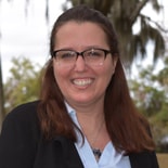 A woman with long brown hair and glasses smiles outside in a black blazer, trees behind her, representing medbridge.