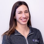 A woman with long brown hair in a gray "TRY" jacket smiles in front of a plain background, reflecting medbridge innovation.