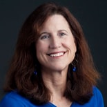 Woman with long brown hair in a blue top, smiling before a dark background, representing medbridge digital healthcare.