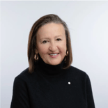 A smiling woman with straight brown hair, a black turtleneck, and gold earrings poses in front of a plain light background.