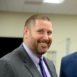 A smiling man in a gray suit with a purple shirt and patterned tie stands indoors, reflecting digital healthcare professionalism.