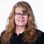 Woman with wavy light brown hair and glasses, wearing a black jacket, smiles in front of a white background.