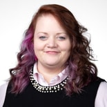 A woman with wavy brown and purple hair, dressed in a black top over a pink collared shirt, smiles at the camera against a white background for medbridge.