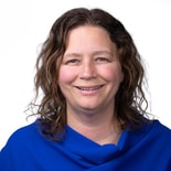 A woman with wavy brown hair in a blue top smiles at the camera, symbolizing hybrid care and medbridge digital health.