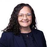 Woman with curly dark hair, glasses, and a navy blazer smiles at the camera against a white background.