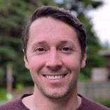 A smiling man with short brown hair and a beard stands outdoors near trees and a blurred medbridge-related building.